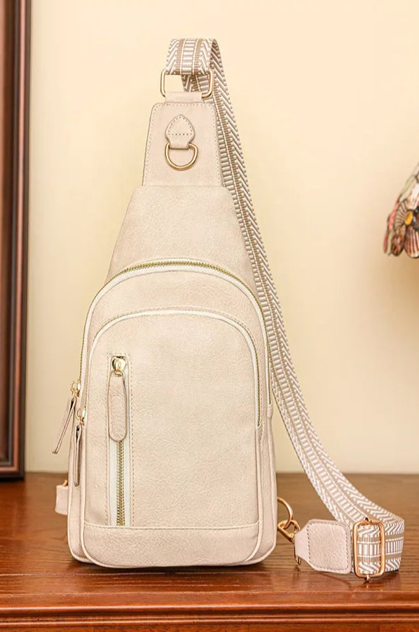 Sac à bandoulière beige clair avec détails dorés, posé sur une commode en bois, style élégant avec sangle textile à motif et fond décoratif « fashion ».

